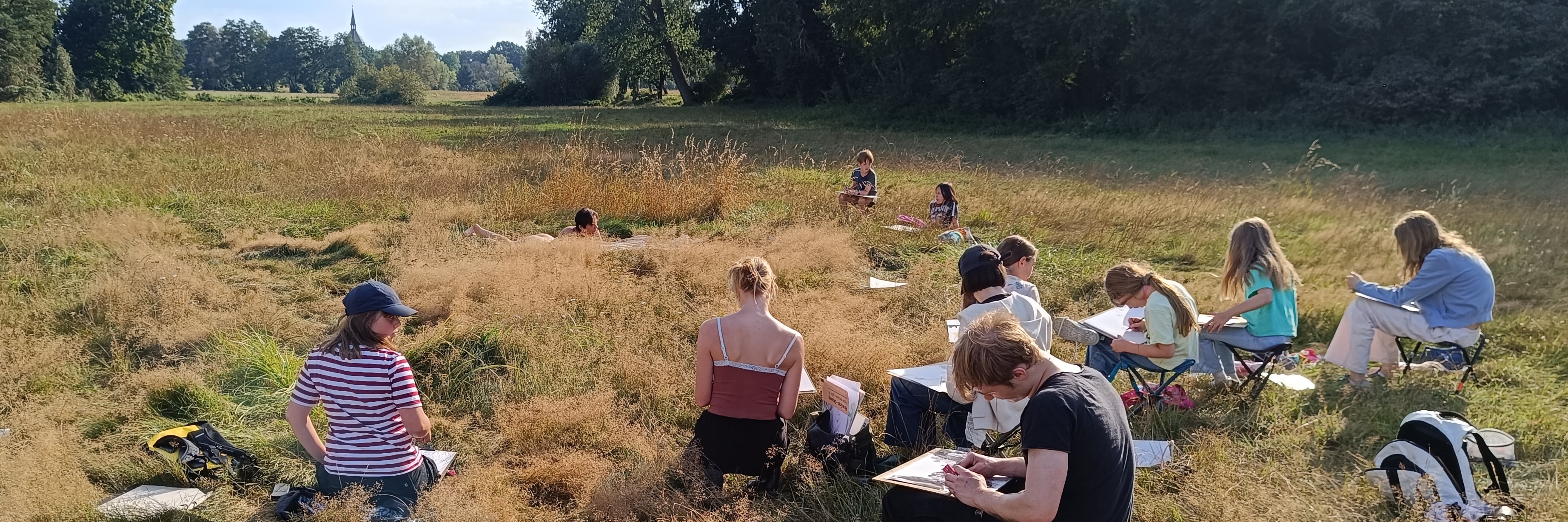 Schüler einer Kunstschule zeichnen gemeinsam in einer weiten Wiesenlandschaft unter freiem Himmel.