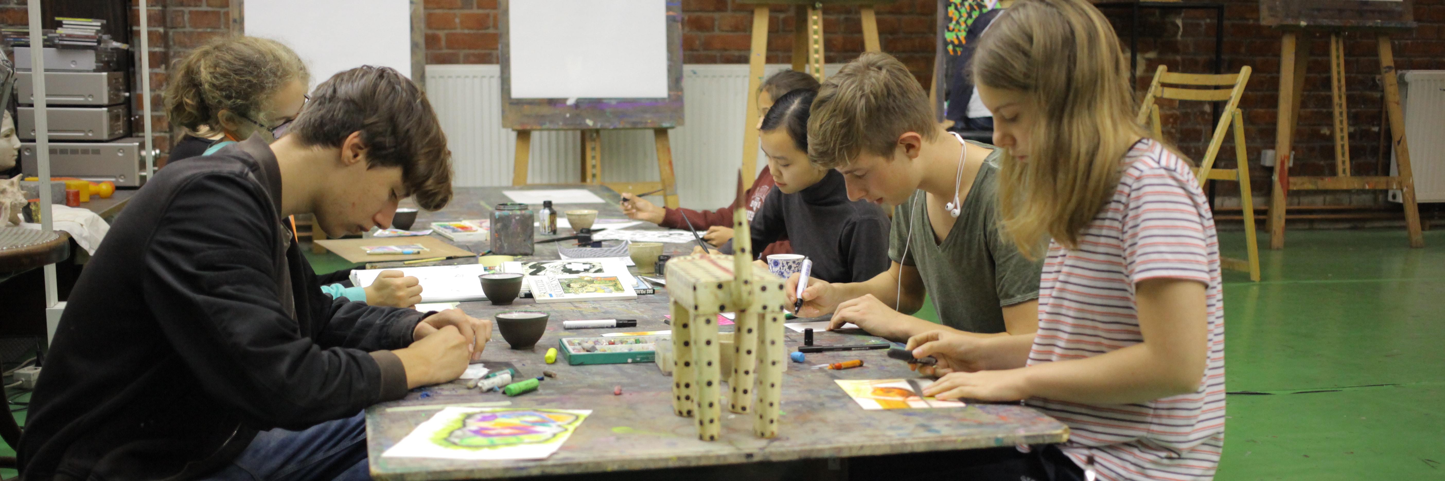 Mehrere Jugendliche zeichnen und malen konzentriert an einem Tisch in einem Kunstschul-Atelier mit Staffeleien im Hintergrund.