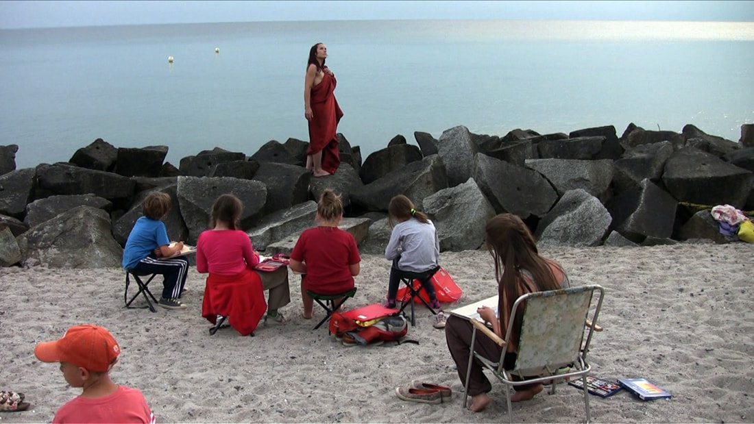 Gruppe zeichnet am Strand nach einem stehenden Modell in rotem Kleid vor dem Meer.