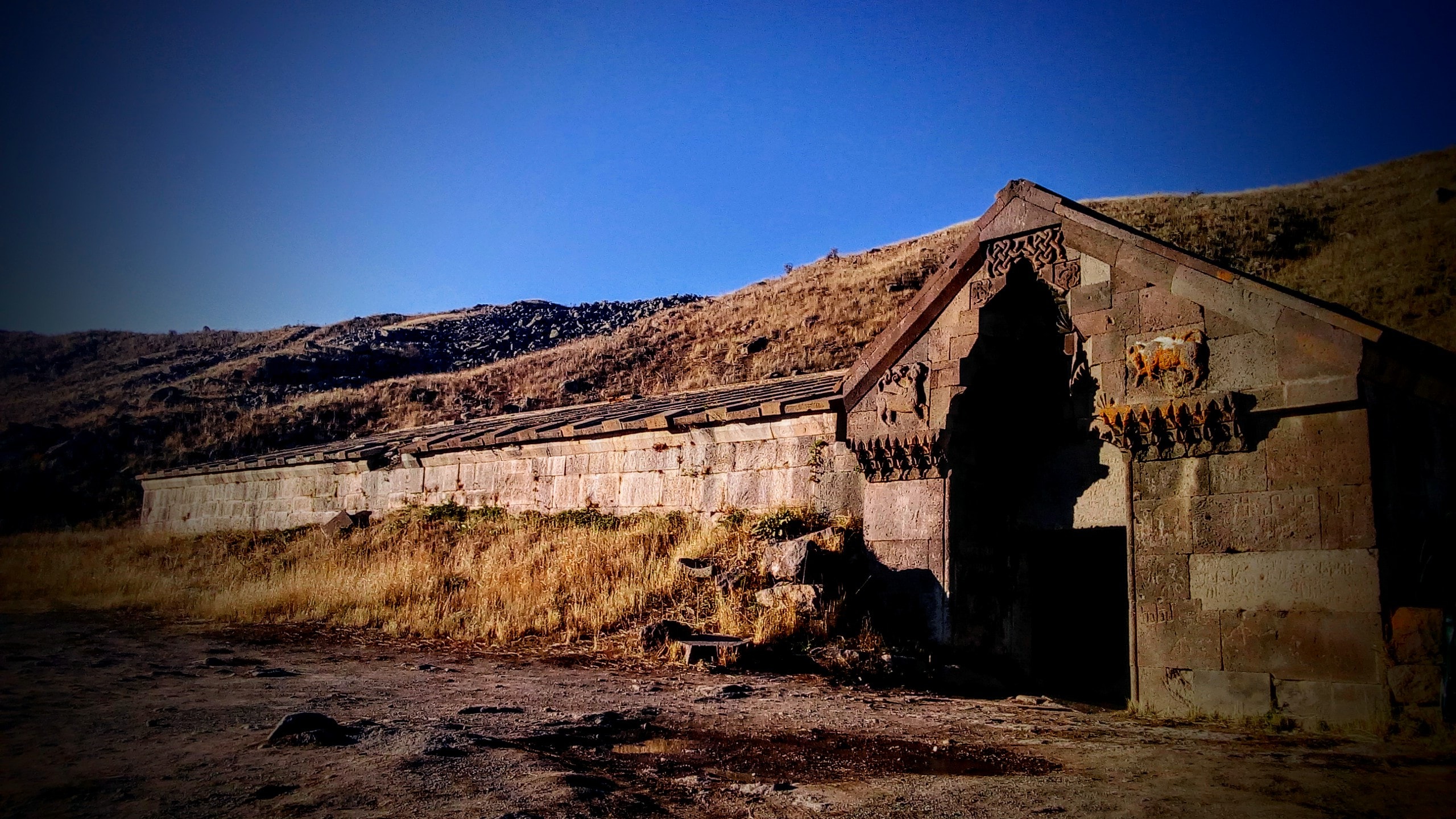 Steinbau mit verziertem Portal und Mauer vor Berghang bei Dämmerlicht.