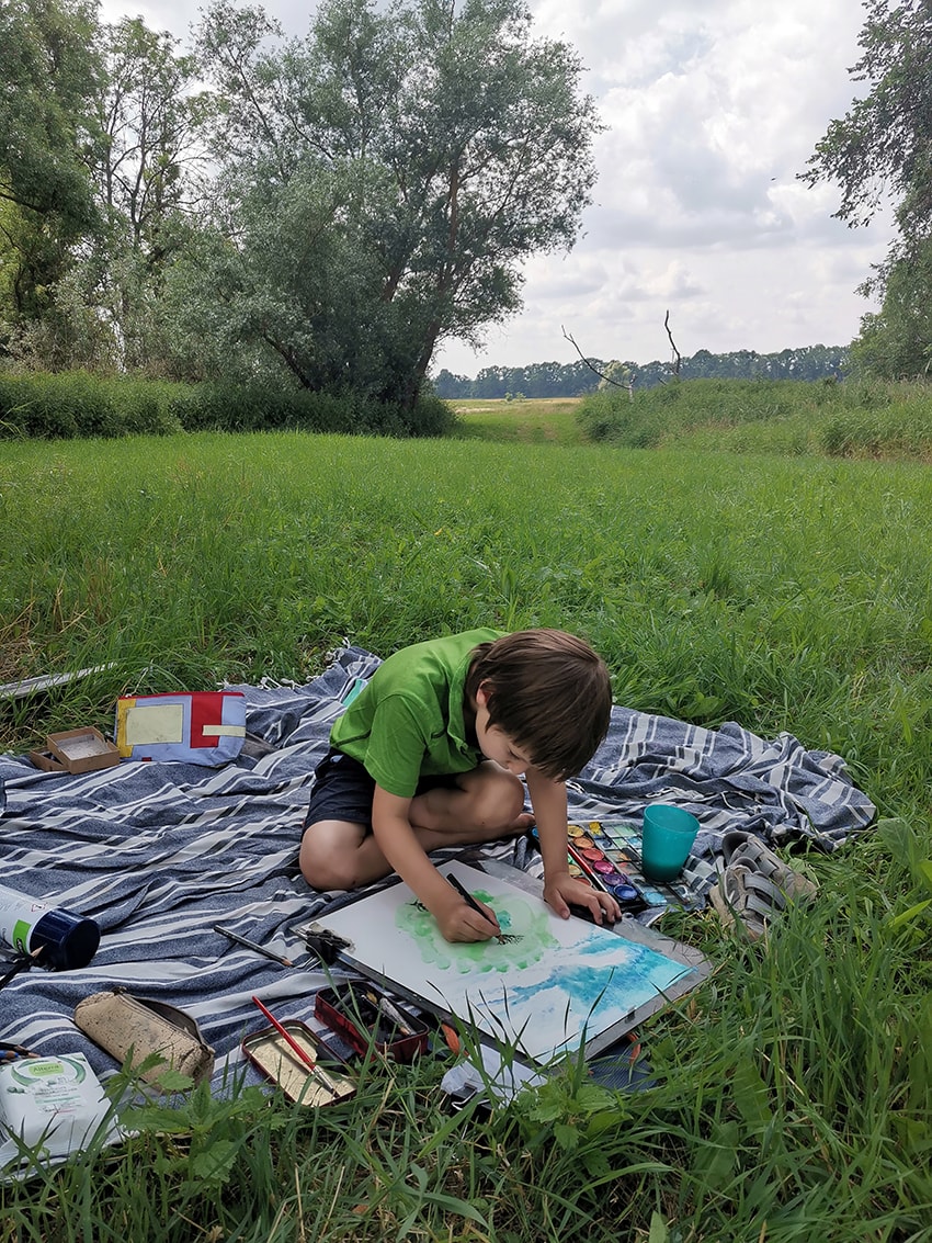 Junge in grünem Hemd malt mit Aquarellfarben auf einer Decke in der Wiese.