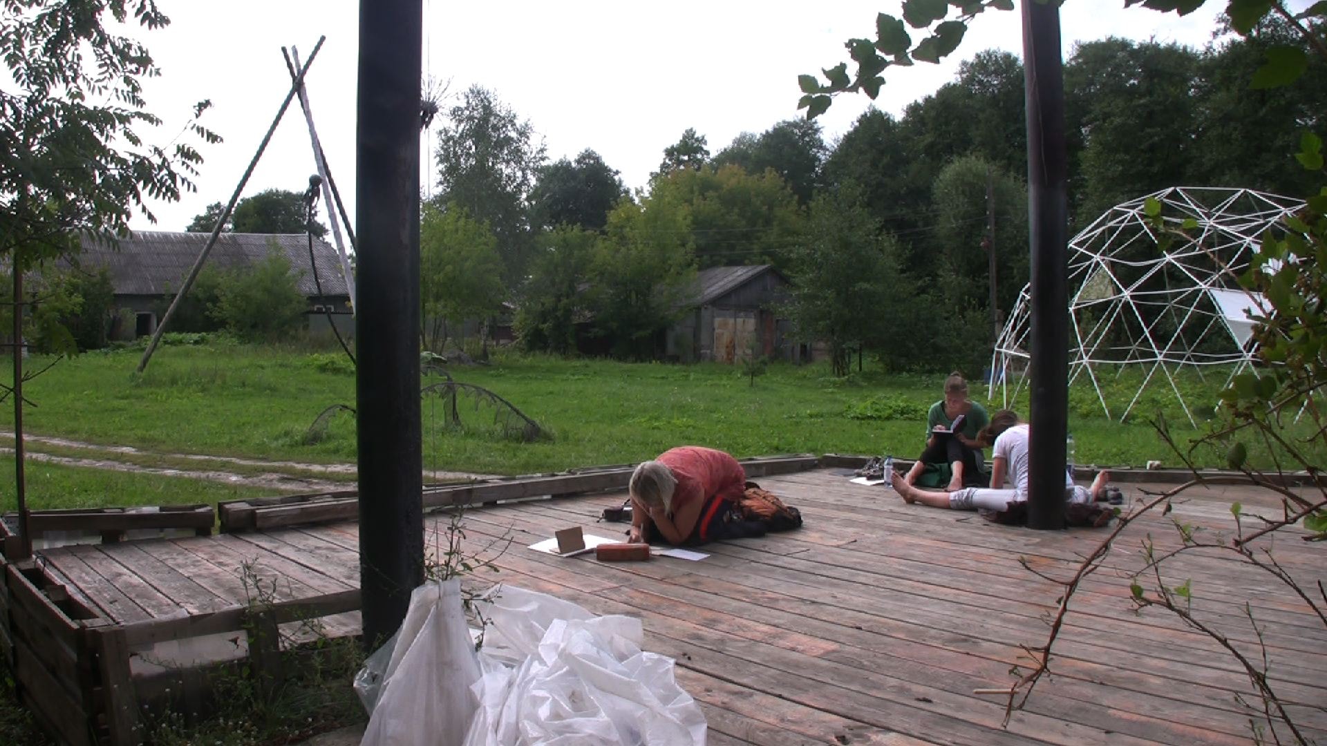 Drei Personen zeichnen auf einer Holzterrasse vor Wiese und Bauernhaus.