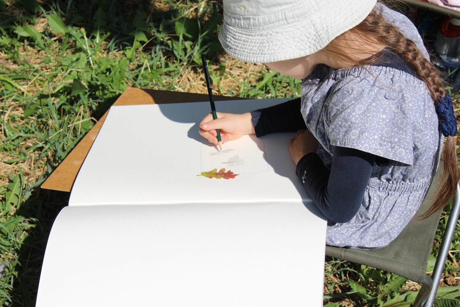Mädchen mit Sonnenhut zeichnet im Gras ein Blatt in ein großes Skizzenbuch.