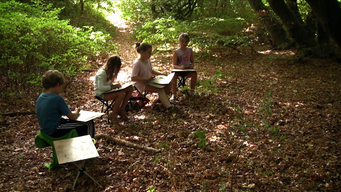 Vier Kinder sitzen verteilt im sonnigen Wald und zeichnen auf Klemmbrettern und Staffeleien.