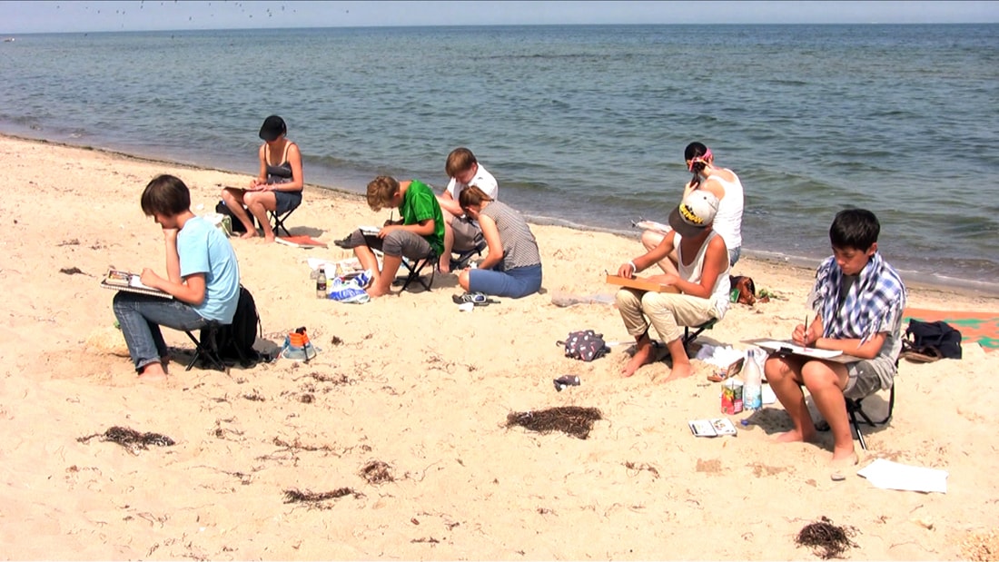 Jugendliche und Erwachsene sitzen am Sandstrand mit Skizzenblöcken und zeichnen, dahinter das Meer.